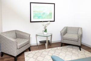 Two patterned armchairs in a bright waiting area with a glass side table, white orchid arrangement, framed artwork, and a neutral area rug.