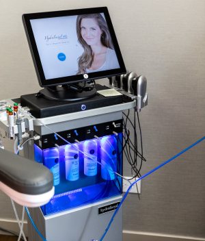 HydraFacial MD treatment device with touchscreen, handpieces, and illuminated solution bottles in a treatment room.