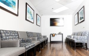 Bright medical office waiting room with patterned gray chairs, wood flooring, framed wall art, and a wall-mounted TV.